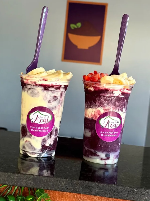Dois copos de açaí com o adesivo da Casa do Açaí, cobertos de banana e morango, com colheres roxas