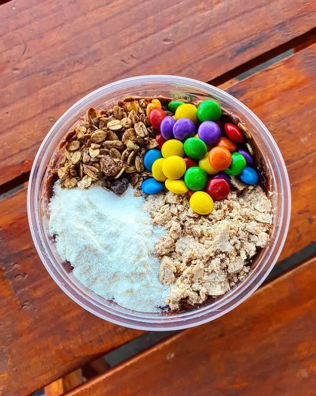 Tigela de açaí vista de cima, dividida em granola, confeitos de chocolate coloridos, leite em pó e paçoca